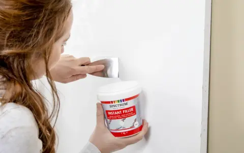 Kobieta używa szpachli i pojemnika z masą szpachlową Spectrum Instant Filler, by czym zaszpachlować dziurę w białej ścianie.
