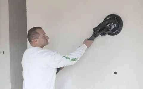 Mężczyzna w okularach ochronnych szlifuje ścianę gładzią z włóknem szklanym, przygotowując ją do malowania.