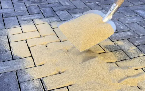 Łopata rozsypuje piasek na kostce brukowej. To kluczowy etap prac, który decyduje, jak długo ma leżeć piasek na kostce brukowej, by zapewnić stabilność.