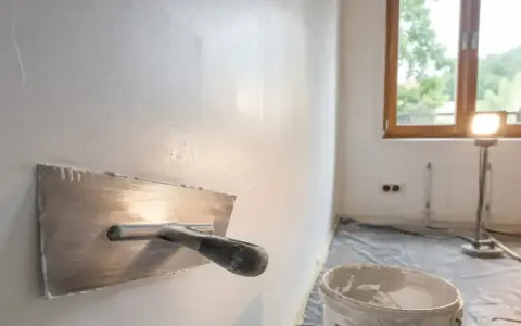 Gips na ścianę jest nakładany kielnią. W tle okno i lampa budowlana.