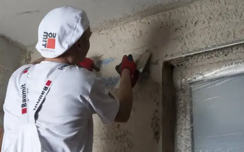 Pracownik w białej czapce i fartuchu wykonuje zacieranie tynku gipsowego na ścianie, używając kielni.