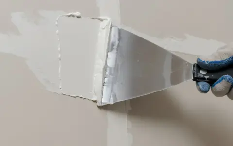 Ręka w rękawicy nakłada masę do łączenia płyt gk na ścianę za pomocą szpachli.