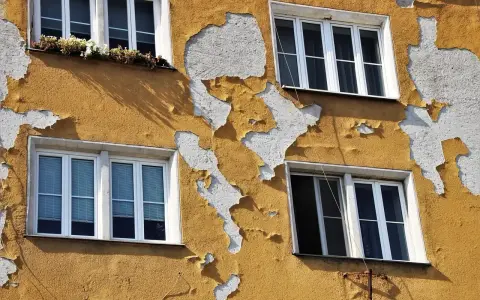 Ściana budynku z żółtym tynkiem baranek, który łuszczy się, odsłaniając biały podkład. Widać cztery okna.