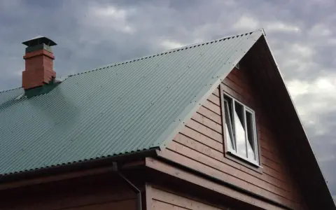 Dach domu pokryty zieloną blachą trapezową, z ceglanym kominem i drewnianą ścianą. Precyzyjne cięcie blachy trapezowej zapewnia estetykę.