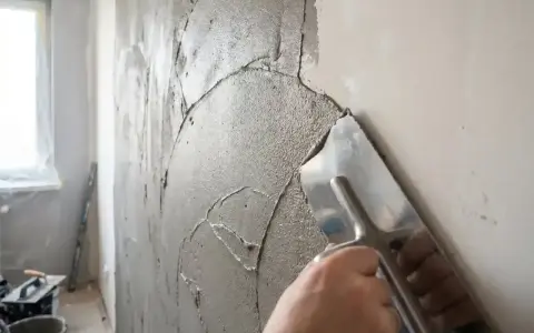 Ręka zacierająca świeżą masę naprawczą do tynku cementowo-wapiennego na ścianie.