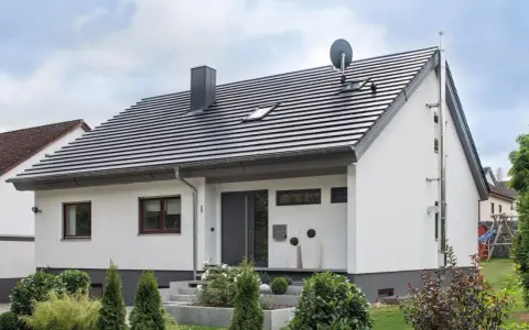 Nowoczesny dom z białymi ścianami i szarym dachem o nachyleniu 45 stopni. Na dachu komin i antena satelitarna.