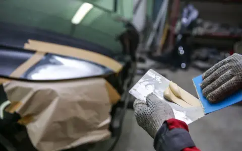 Ręce w rękawiczkach nakładają szpachlę elastyczną na pęknięcia karoserii samochodu.
