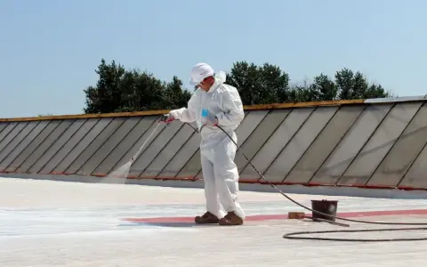Pracownik w białym kombinezonie natryskuje białą masę na płaski dach, szukając alternatywy dla papy.