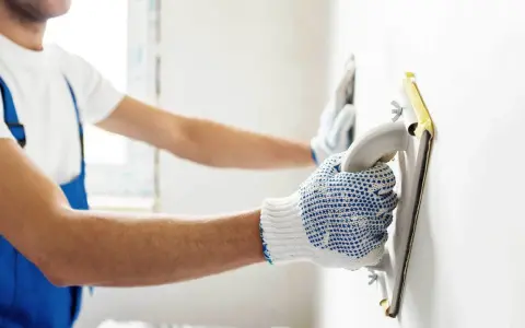 Pracownik w niebieskim ogrodniczkach wygładza ścianę pacą. Zastanawiasz się, ile gładzi na m2 zużyjesz?