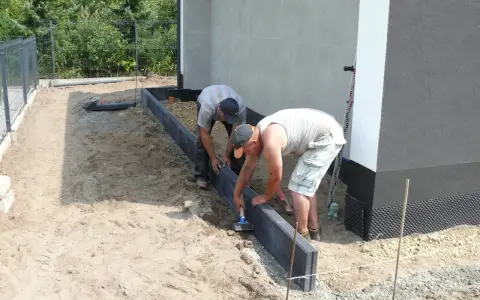 Dwóch mężczyzn zajmuje się układaniem obrzeży betonowych wokół budynku.