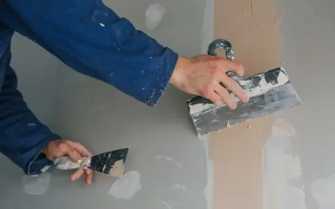 Ręce pracownika w niebieskim kombinezonie nakładają gładź szpachlową na ścianę. Dowiedz się, jak przyspieszyć schnięcie gładzi.