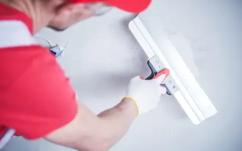 Ręce w białych rękawiczkach z czerwonymi akcentami wygładzają ścianę za pomocą szerokiej kielni. Gładź na mokro tworzy jednolitą powierzchnię.