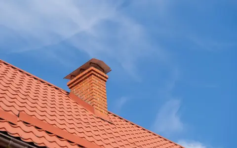 Obróbka komina blachą modułową na dachu z czerwonej dachówki. Niebo błękitne z delikatnymi chmurkami.