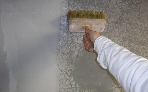 Ręka w białej bluzie nakłada preparat pędzlem na popękaną gładź, by ją naprawić.