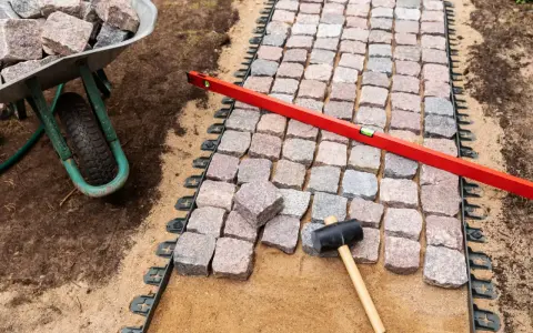 Układanie kostki brukowej z progiem. Wózek z kamieniami, poziomica i gumowy młotek czekają na dokończenie ścieżki.