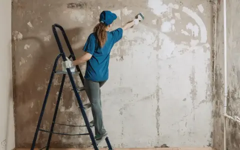 Kobieta na drabinie usuwa gładź ze ściany za pomocą pędzla. Proces przygotowania do remontu.