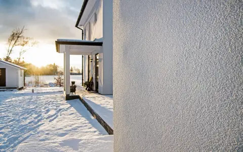 Białe, chropowate tynki na ścianach domu w zimowy, słoneczny dzień. Śnieg pokrywa podwórko i otoczenie.