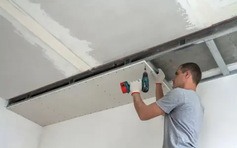 Mężczyzna przykręca płytę kartonowo-gipsową do stelaża sufitowego. Do cięcia karton gips używa się specjalnych narzędzi.