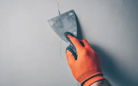 Ręka w pomarańczowej rękawicy zacierająca pionowe pęknięcia ścian działowych metalową szpachlą.
