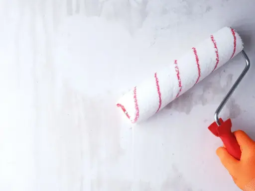 Ręka w pomarańczowej rękawicy gruntuje zeszklona ścianę wałkiem z czerwonymi paskami.
