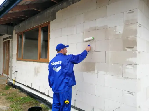 Pracownik w niebieskim kombinezonie wykonuje gruntowanie ścian z bloczków przed tynkowaniem, używając wałka.