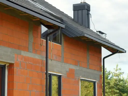 Nowy dom w budowie, z widocznymi cegłami i oknami. Zastanawiasz się, jakie rury do odprowadzenia wody z rynien wybrać? Tutaj widzisz czarne rury spustowe.