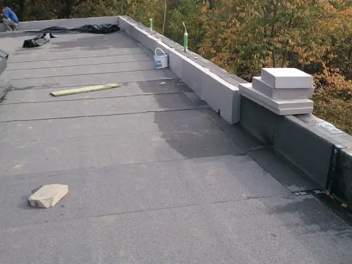 Prace dekarskie: układanie papy na stromym dachu. Widać materiały budowlane i pracowników.