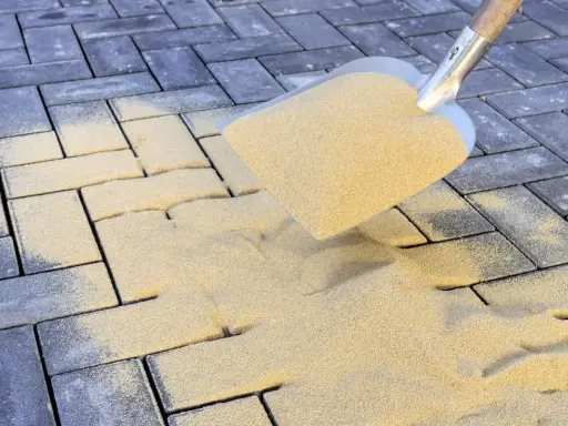Łopata rozsypuje piasek na kostce brukowej. To kluczowy etap prac, który decyduje, jak długo ma leżeć piasek na kostce brukowej, by zapewnić stabilność.