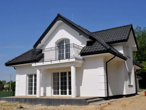 Nowy dom z białym gipsem na zewnątrz, czarnym dachem i balkonem. Wokół plac budowy z piaskiem.