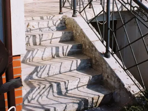 Kamienne stopnie schodów zewnętrznych, z widocznymi cieniami balustrady. Jaki beton na schody zewnętrzne? Ten wygląda na solidny.