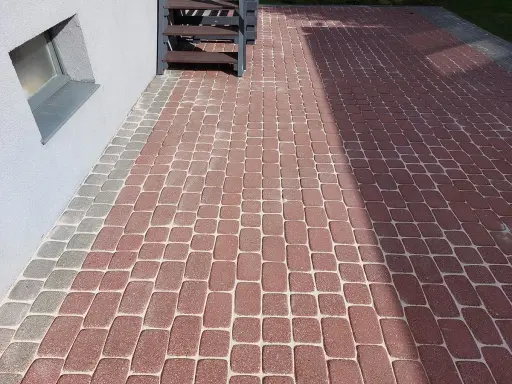 Ubijanie kostki z piaskiem czy bez? Świeżo ułożona ścieżka z czerwonej i szarej kostki brukowej przy domu, z drewnianymi schodkami.