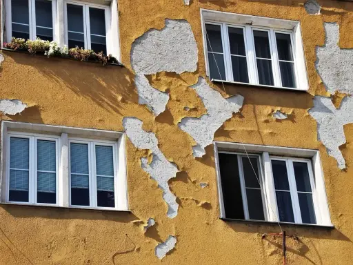 Ściana budynku z żółtym tynkiem baranek, który łuszczy się, odsłaniając biały podkład. Widać cztery okna.