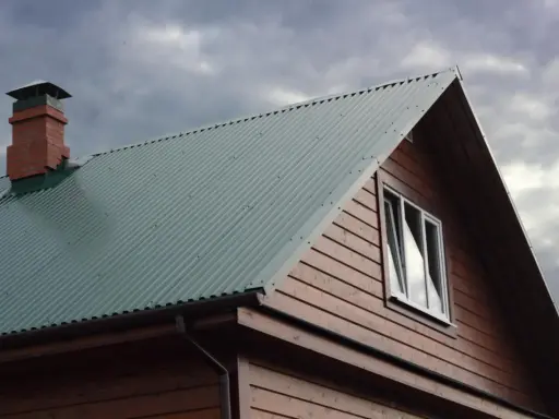 Dach domu pokryty zieloną blachą trapezową, z ceglanym kominem i drewnianą ścianą. Precyzyjne cięcie blachy trapezowej zapewnia estetykę.