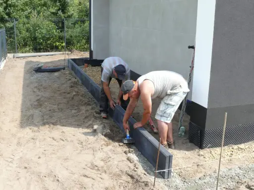 Dwóch mężczyzn zajmuje się układaniem obrzeży betonowych wokół budynku.