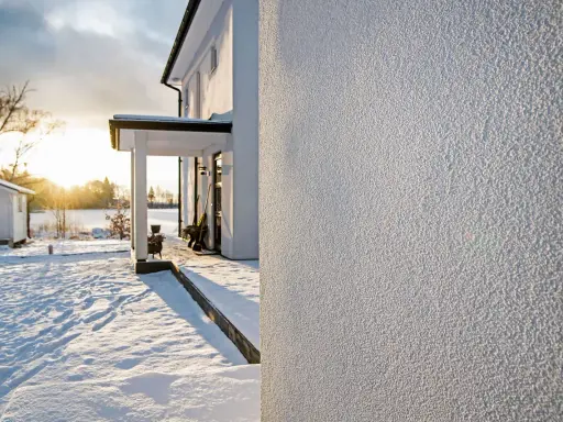 Białe, chropowate tynki na ścianach domu w zimowy, słoneczny dzień. Śnieg pokrywa podwórko i otoczenie.