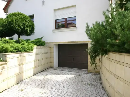 Wjazd z kostki brukowej prowadzi do ciemnobrązowej bramy garażowej. Otaczają go kamienne murki z bujną zielenią.