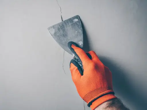 Ręka w pomarańczowej rękawicy zacierająca pionowe pęknięcia ścian działowych metalową szpachlą.