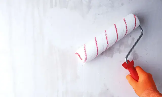 Ręka w pomarańczowej rękawicy gruntuje zeszklona ścianę wałkiem z czerwonymi paskami.