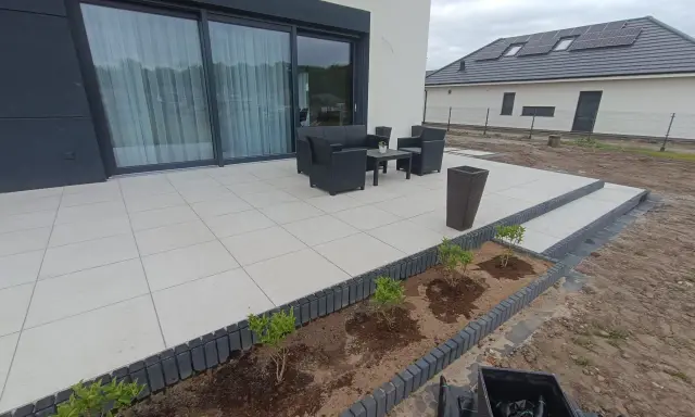 Nowy taras z płyt betonowych czeka na meble ogrodowe. Wokół rabaty z młodymi krzewami.