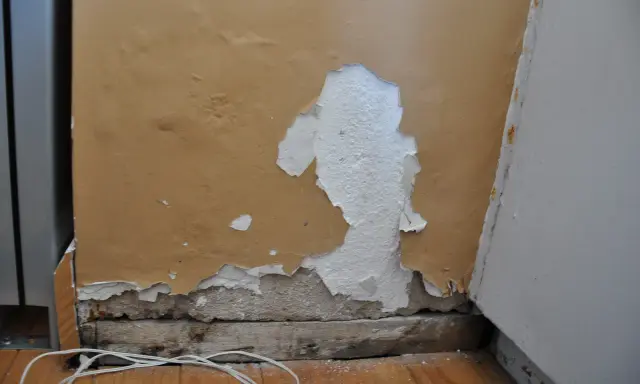 Zła gładź na ścianie, łuszcząca się farba odsłania tynk. Widoczne drewniane belki i biały kabel na podłodze.