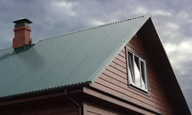 Dach domu pokryty zieloną blachą trapezową, z ceglanym kominem i drewnianą ścianą. Precyzyjne cięcie blachy trapezowej zapewnia estetykę.