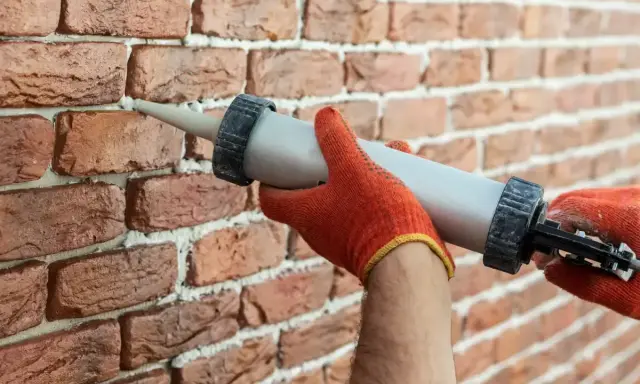 Ręce w pomarańczowych rękawicach używają pistoletu z masą uszczelniającą, by wypełnić ubytki w cegle.