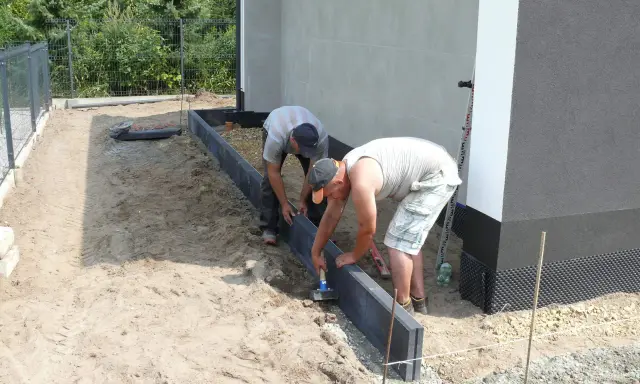 Dwóch mężczyzn zajmuje się układaniem obrzeży betonowych wokół budynku.