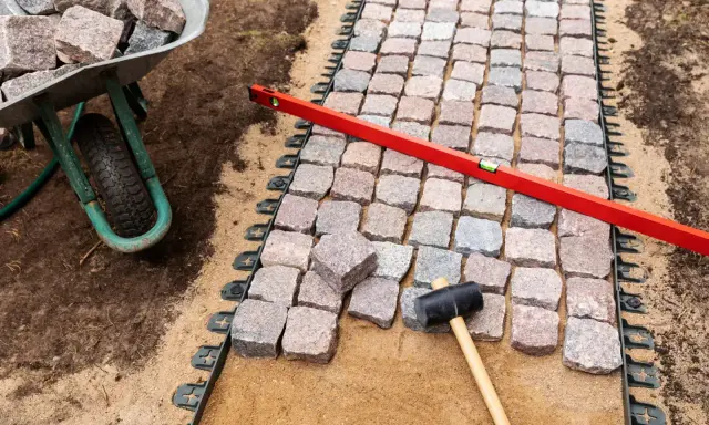 Układanie kostki brukowej z progiem. Wózek z kamieniami, poziomica i gumowy młotek czekają na dokończenie ścieżki.