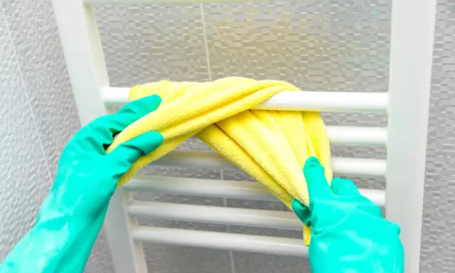 Ręce w zielonych rękawiczkach czyszczą biały grzejnik żółtą ściereczką. Dowiedz się, jak wyczyścić grzejnik z kurzu w środku, aby zapewnić higienę.