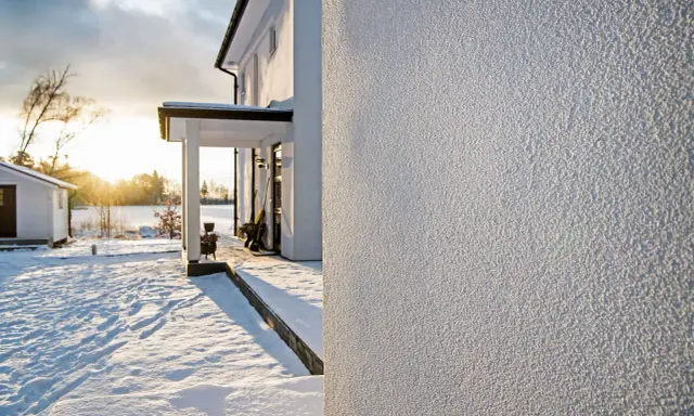 Białe, chropowate tynki na ścianach domu w zimowy, słoneczny dzień. Śnieg pokrywa podwórko i otoczenie.