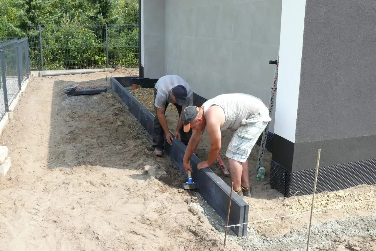 Dw&oacute;ch mężczyzn pracuje przy układaniu obrzeży betonowych wok&oacute;ł budynku.
