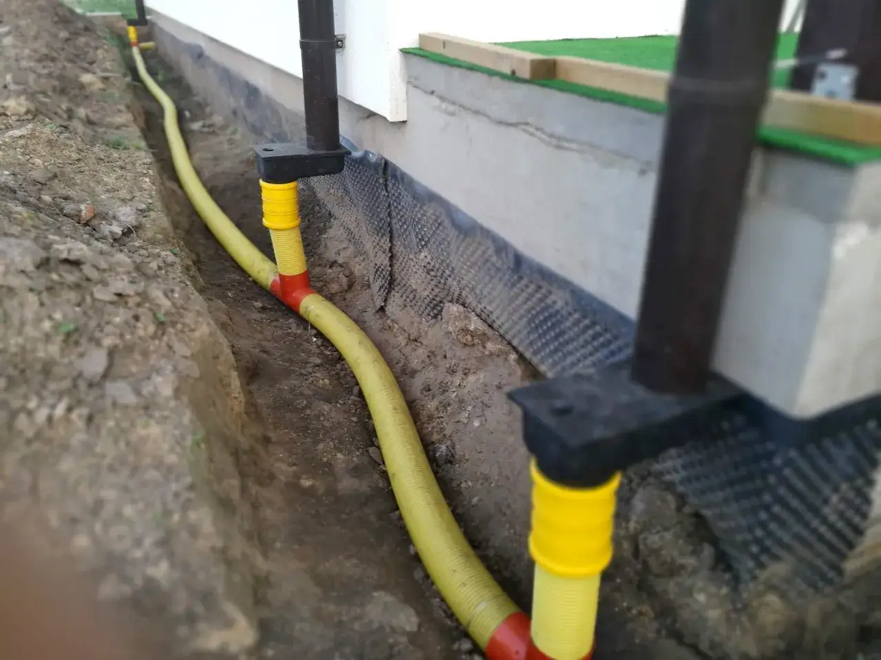 Instalacja odprowadzająca wodę z rynien, zgodna z przepisami, z ż&oacute;łtą rurą biegnącą w wykopie.