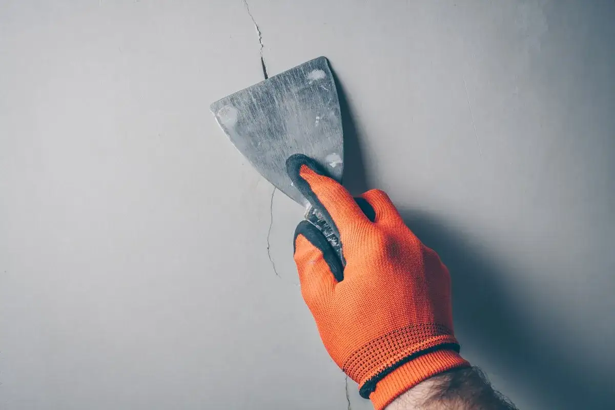 Ręka w pomarańczowej rękawicy próbuje naprawić pęknięcie na ścianie, ale widać, że źle położona gładź nie ukryje defektu.