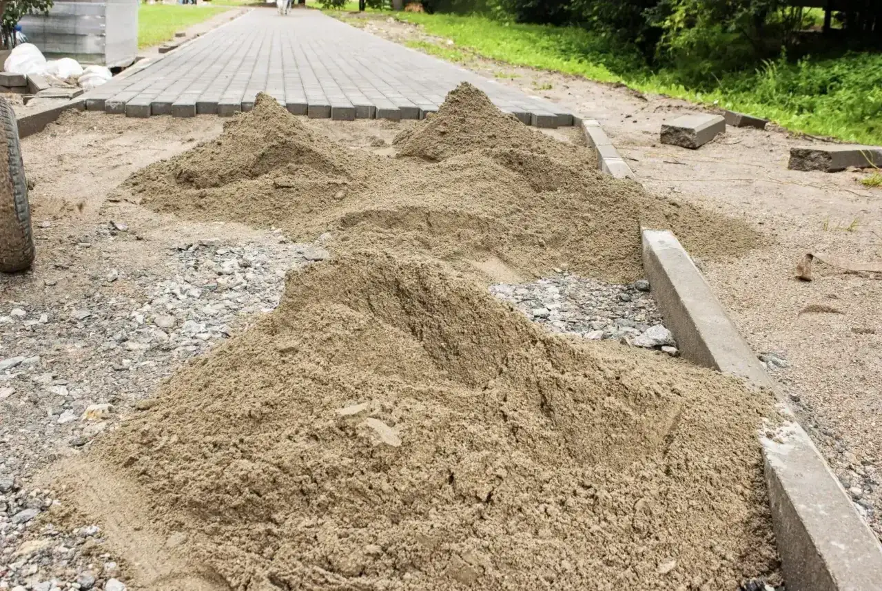 Na placu budowy widać kopce piasku, kostkę brukową i krawężniki. Trwa ubijanie kostki z piaskiem, przygotowując ścieżkę.